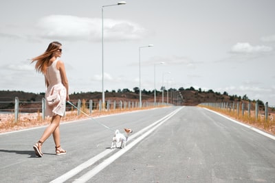 白天，一名身穿白色无袖连衣裙的妇女牵着小狗的背带在柏油路上行走